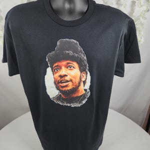 Pu&ograve; includere: T-shirt nero con un ritratto di un uomo con la barba e un cappello di pelliccia. Il ritratto &egrave; in un cerchio bianco. La t-shirt &egrave; esposta su un manichino nero.
