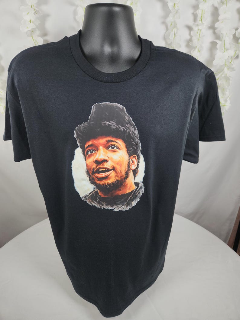 Pu&ograve; includere: T-shirt nero con un ritratto di un uomo con la barba e un cappello di pelliccia. Il ritratto &egrave; in un cerchio bianco. La t-shirt &egrave; esposta su un manichino nero.