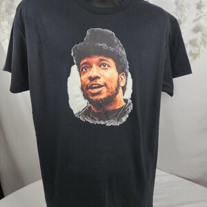 Pu&ograve; includere: T-shirt nero con un ritratto di un uomo con capelli scuri e barba. Il ritratto &egrave; ambientato su uno sfondo bianco. La t-shirt &egrave; esposta su un manichino nero.