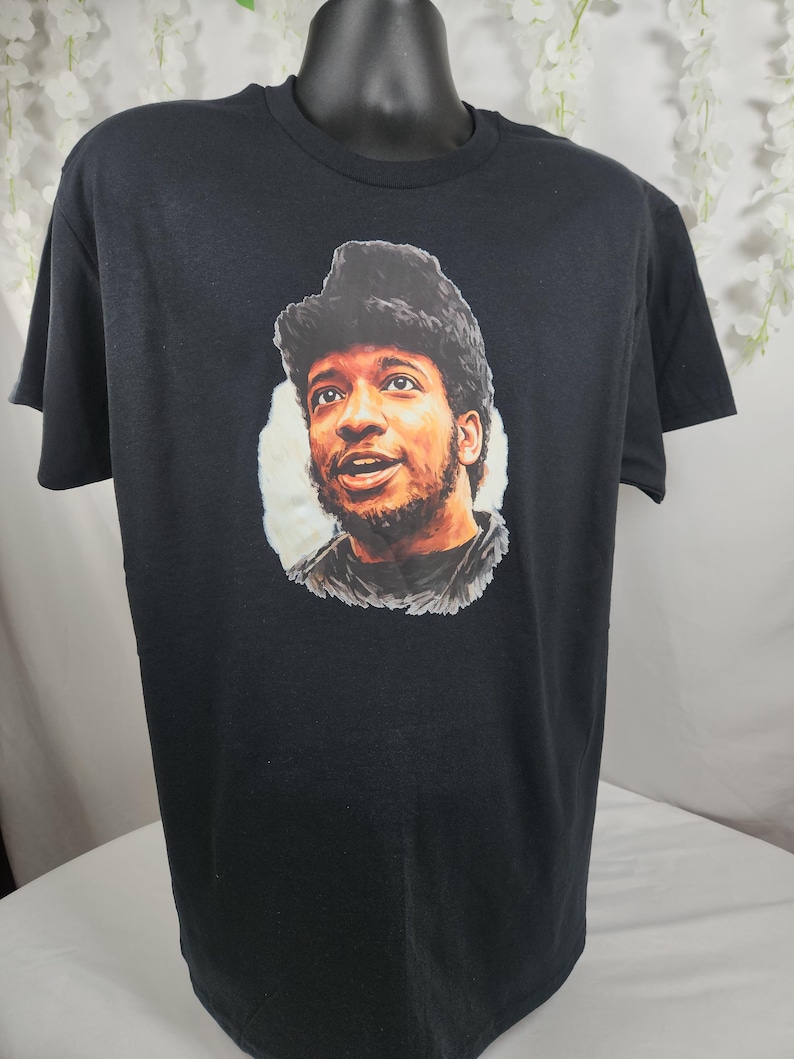 Pu&ograve; includere: T-shirt nero con un ritratto di un uomo con capelli scuri e barba. Il ritratto &egrave; ambientato su uno sfondo bianco. La t-shirt &egrave; esposta su un manichino nero.