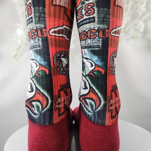 May include: Pair of red and orange socks featuring the text "NCCU Eagles" and an eagle graphic. The socks have a maroon toe and heel. The design includes the words "North Carolina Central University".