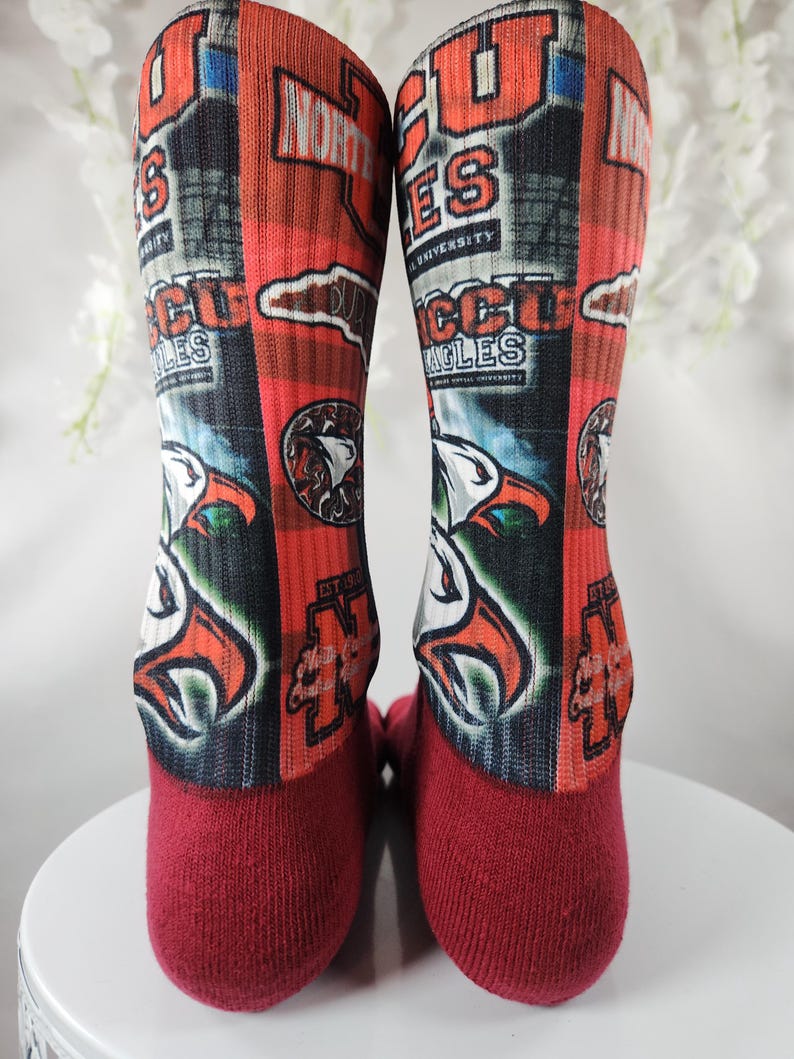 May include: Pair of red and orange socks featuring the text "NCCU Eagles" and an eagle graphic. The socks have a maroon toe and heel. The design includes the words "North Carolina Central University".