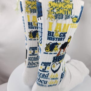 Può includere: Un paio di calzini bianchi con testo e grafica gialli e blu. Il testo include "I AM BLACK HISTORY", "Johnson C. Smith University" e "HBCU". I calzini hanno un simbolo di pace e un cuore.