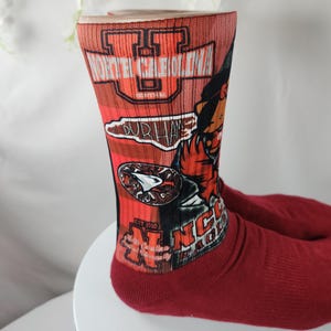 May include: A pair of red socks with a graphic design featuring the text "NORTH CAROLINA" and "DURHAM". The socks also display a stylized image of a baseball player and the text "NC".