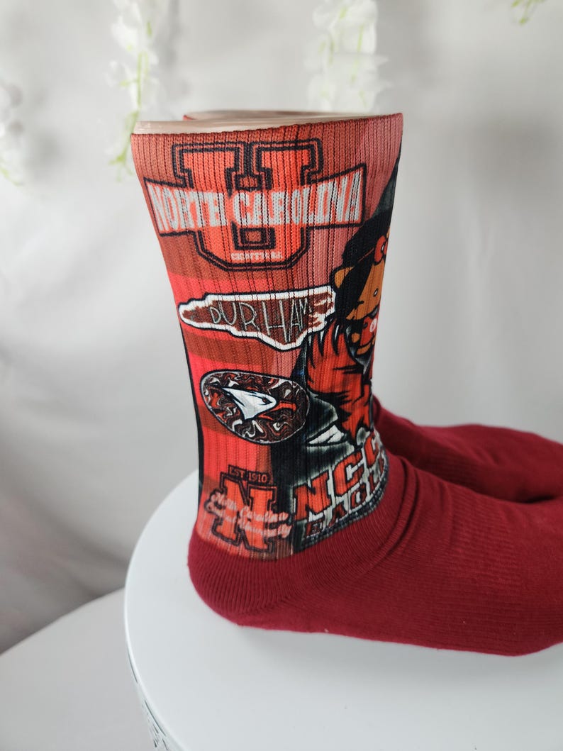 May include: A pair of red socks with a graphic design featuring the text "NORTH CAROLINA" and "DURHAM". The socks also display a stylized image of a baseball player and the text "NC".