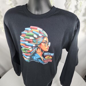 以下が含まれることがあります： 黒い長袖Tシャツ。髪に本をつけた女性のカラフルなグラフィックが特徴で、メガネと大きなフープイヤリングを着用しています。「EDUCATION IS FREEDOM」の文字がプリントされています。Tシャツは黒いマネキンに展示されています。