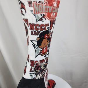 May include: A white crew sock featuring a repeating graphic print of North Carolina Central University branding, including the text NCCU EAGLES, an eagle mascot, and a cartoon character wearing a red jersey with a bow, with a black heel and toe.