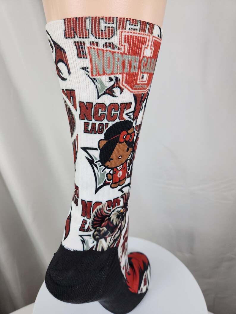 May include: A white crew sock featuring a repeating graphic print of North Carolina Central University branding, including the text NCCU EAGLES, an eagle mascot, and a cartoon character wearing a red jersey with a bow, with a black heel and toe.