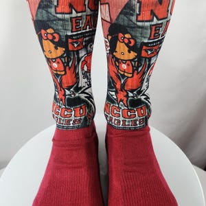 May include: Pair of red socks with a design featuring the letters "NCCU EAGLES" in orange and white. The socks have a cartoon eagle mascot and the words "NORTH CAROLINA" on them.
