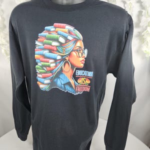 以下が含まれることがあります： 黒い長袖Tシャツ。髪に本をつけた女性のカラフルなグラフィックが特徴で、メガネと金のフープイヤリングを着用しています。「EDUCATION IS FREEDOM」の文字が画像の下に印刷されています。マネキンにシャツが表示されています。