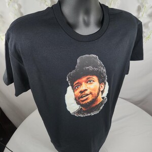 Pu&ograve; includere: T-shirt nero girocollo con un ritratto di un uomo con un cappello di pelliccia. Il ritratto &egrave; in una cornice circolare. La t-shirt &egrave; esposta su un manichino nero.