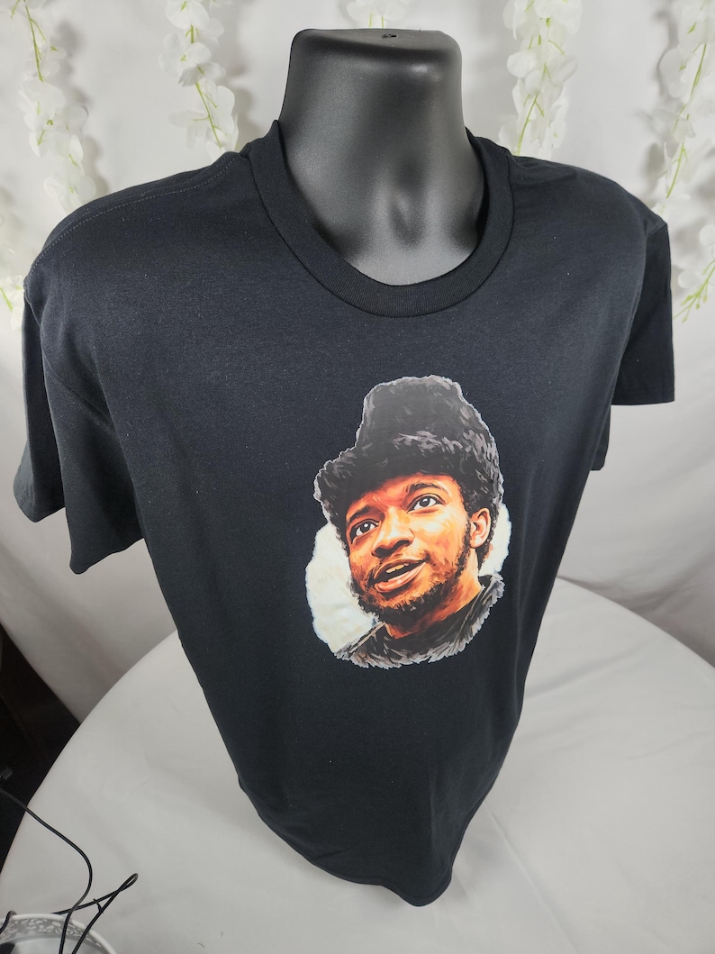 Pu&ograve; includere: T-shirt nero girocollo con un ritratto di un uomo con un cappello di pelliccia. Il ritratto &egrave; in una cornice circolare. La t-shirt &egrave; esposta su un manichino nero.