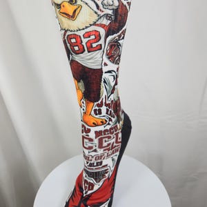 May include: A white crew sock featuring a graphic of a bald eagle mascot wearing a jersey with the number 82. The design includes red and black accents, text reading NCCU SCHOOL OF LAW, and additional athletic-themed illustrations throughout.