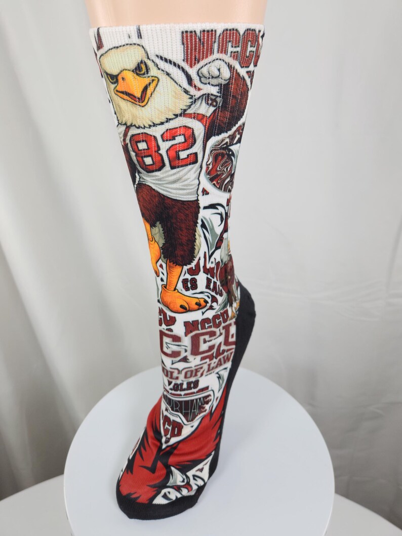 May include: A white crew sock featuring a graphic of a bald eagle mascot wearing a jersey with the number 82. The design includes red and black accents, text reading NCCU SCHOOL OF LAW, and additional athletic-themed illustrations throughout.