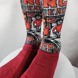 May include: A pair of red socks with a design featuring the text "NC EAGLES" in orange and white, along with a cartoon eagle mascot. The socks have a ribbed cuff and a detailed graphic.