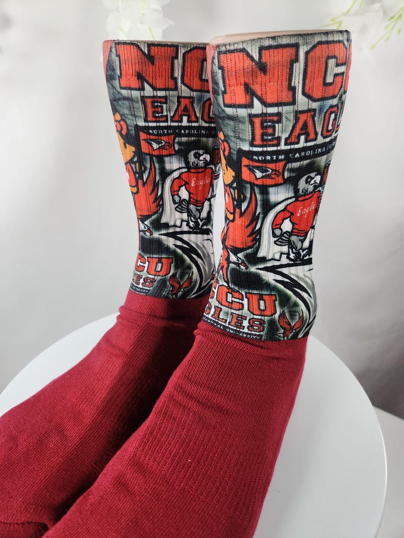 May include: A pair of red socks with a design featuring the text "NC EAGLES" in orange and white, along with a cartoon eagle mascot. The socks have a ribbed cuff and a detailed graphic.
