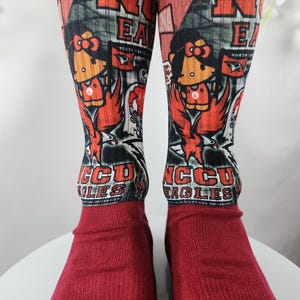 May include: A pair of red socks with a graphic design featuring the letters "NCCU EAGLES" and a cartoon character. The socks have a red foot and a patterned upper section with the team's logo and mascot.