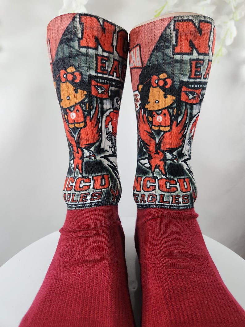 May include: A pair of red socks with a graphic design featuring the letters "NCCU EAGLES" and a cartoon character. The socks have a red foot and a patterned upper section with the team's logo and mascot.
