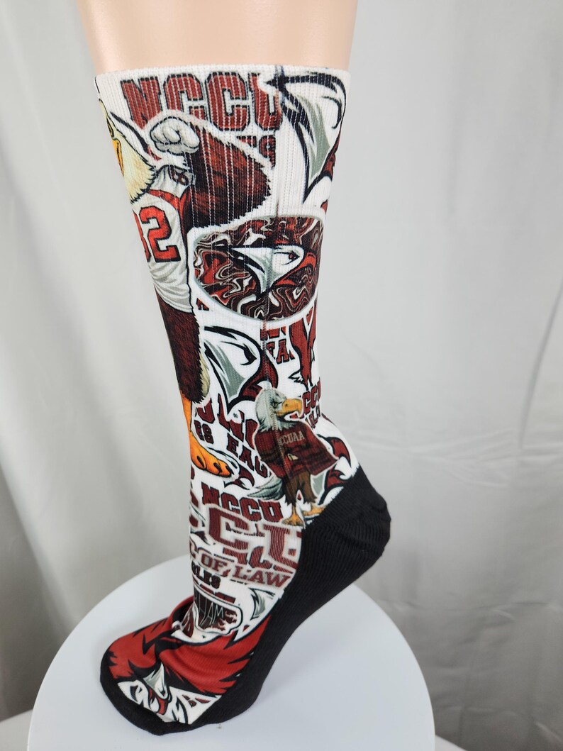 May include: A white crew sock featuring a repeating graphic print of the NCCU Eagles mascot, the number 12, and the text NCCU. The design includes red, black, and white colors, with a solid black heel and toe section, showcasing various eagle illustrations.