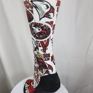 May include: A white crew sock featuring an all-over print of eagle graphics in red, brown, and white, along with the text 'NCCU EAGLES'. The sock has a solid black heel and toe section, with a ribbed texture extending from the ankle to the calf.