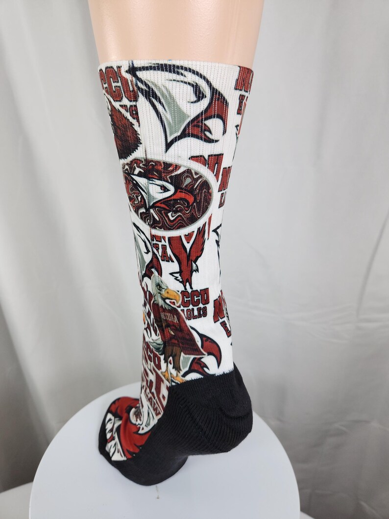 May include: A white crew sock featuring an all-over print of eagle graphics in red, brown, and white, along with the text 'NCCU EAGLES'. The sock has a solid black heel and toe section, with a ribbed texture extending from the ankle to the calf.
