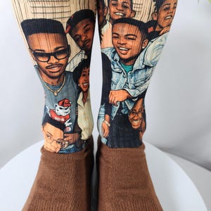 May include: A pair of brown socks with a ribbed texture on the foot and a printed design on the upper portion. The design features a group of smiling people in a cartoon style. The socks are displayed on a white surface.