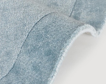 May include: A light blue area rug with a dense, short-pile texture and a curved, folded edge. The rug features a smooth, uniform surface with a subtle sheen, showcasing a consistent cool-toned blue color throughout the visible fabric material.