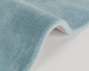 May include: Close-up of a light blue rug with a soft, plush texture. The rug is folded over, revealing a glimpse of the underside. The color is a muted, pastel blue, creating a calming aesthetic. The rug appears to be made of a dense, woven material.