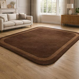 May include: A rectangular brown rug with a lighter brown border. The rug is placed on a hardwood floor in a living room setting. The rug has rounded corners and a simple, elegant design.