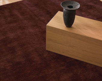 May include: A burgundy rug with a textured surface. A light-colored wooden block is on the rug, with a dark, vase-like object on top. The rug is rectangular and appears to be made of a soft material. The scene is lit with natural light.
