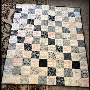 May include: A handmade quilt with a patchwork design. The quilt features a variety of fabrics in shades of blue, white, and black, arranged in a grid pattern. Each square contains handwritten messages and drawings. The quilt is bordered with a black fabric.