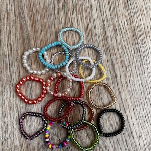 Könnte beinhalten: Eine Sammlung von Perlenringen in verschiedenen Farben, darunter Rot, Schwarz, Gelb und Blau. Einige Ringe haben Perlen, während andere eine Mischung aus bunten Perlen aufweisen. Die Ringe sind auf einer Holzoberfläche angeordnet.