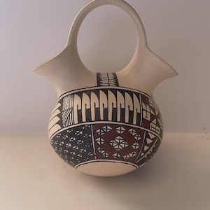 May include: A cream-colored ceramic basket with a rounded base and a handle. The basket is decorated with geometric patterns in black, brown, and white. The handle is a simple arch shape, and the rim flares outward.
