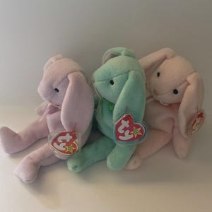 May include: Three pastel-coloured plush bunny toys: pink, light green, and another pink. Each bunny has long ears, a small bow, and a tag. The bunnies are sitting together, creating a soft and cuddly appearance.