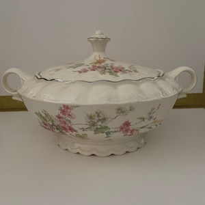 Peut inclure: Plat de service crème avec couvercle et motif floral. Le plat a un bord festonné et deux poignées. Le couvercle est surmonté d'un petit bouton. Le motif floral comprend des fleurs roses, vertes et jaunes.
