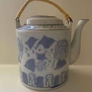 May include: A white and blue ceramic teapot with a floral design. The teapot has a curved spout and a woven handle. The design features blue floral patterns and decorative borders. The teapot is a traditional style.
