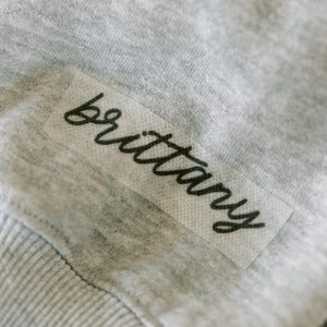 May include: Close-up of a gray fabric with a rectangular label. The label features the name "Brittany" in a cursive, black font. The fabric appears to be a soft, knit material.