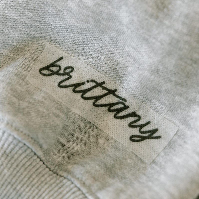 May include: Close-up of a gray fabric with a rectangular label. The label features the name "Brittany" in a cursive, black font. The fabric appears to be a soft, knit material.