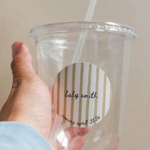 Pode incluir: Um copo descartável de plástico transparente com tampa e palhinha, apresentando um autocolante circular com riscas verticais douradas. O autocolante exibe o texto "baby smith" no centro e "coming april 2026" escrito em cursivo na base.