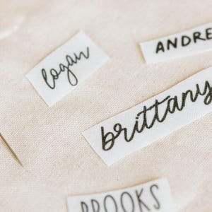 May include: Close-up of fabric name tags with black handwritten names: Logan, Andrew, Brittany, and Brooks. A needle is placed on the light beige fabric. The tags are rectangular and made of white fabric.