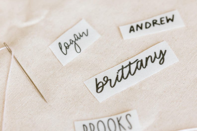 May include: Close-up of fabric name tags with black handwritten names: Logan, Andrew, Brittany, and Brooks. A needle is placed on the light beige fabric. The tags are rectangular and made of white fabric.