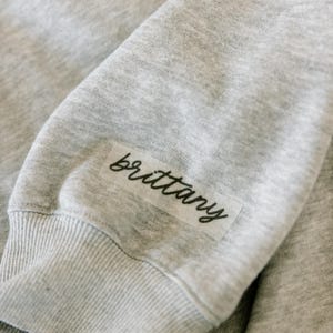 May include: Close-up of a light gray sweatshirt sleeve. A rectangular label with the name "brittany" in black script is affixed to the sleeve near the cuff. The fabric has a soft, textured appearance.