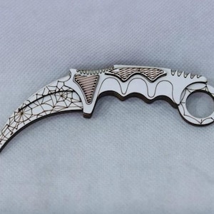 May include: A white and brown karambit knife with a curved blade and finger ring. The blade features a spiderweb design, and the handle has a curved, ergonomic shape. The knife is made of wood and has a decorative appearance.