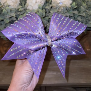 Op de afbeelding: Een grote, paarse satijnen haarstrik versierd met rijen sprankelende strass-steentjes. De strik wordt bij elkaar gehouden door een met strass-steentjes bezette band. De strik heeft een levendige paarse tint, geschikt voor cheerleading of dans.