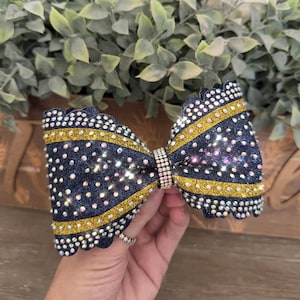 Op de afbeelding: Een haarstrik met een donkerblauwe glitterbasis en geschulpte randen. De strik is geaccentueerd met twee horizontale strepen van goudglitter en bedekt met heldere, reflecterende strass-steentjes. Een centrale band van kleine strass-steentjes houdt de strik vast.