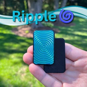 May include: A teal and black rectangular object with a spiral design is held in a hand. The word "Ripple" is written in blue, with a blue spiral graphic. The background is a blurred outdoor scene.