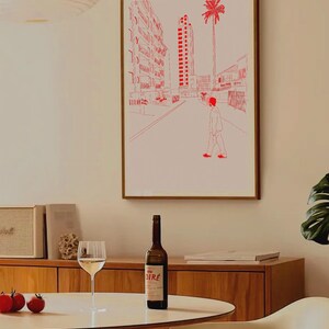 Può includere: Una stampa artistica incorniciata con un disegno a tratto rosso di una scena di strada cittadina con una persona che cammina. Una bottiglia di vino con etichetta è su un tavolo bianco, insieme a un bicchiere da vino. La stanza ha mobili in legno.