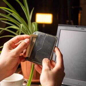 May include: A dark gray leather wallet with white stitching, holding a U.S. dollar bill. The wallet has multiple card slots and is held in front of a laptop. The background includes a plant and a lamp.