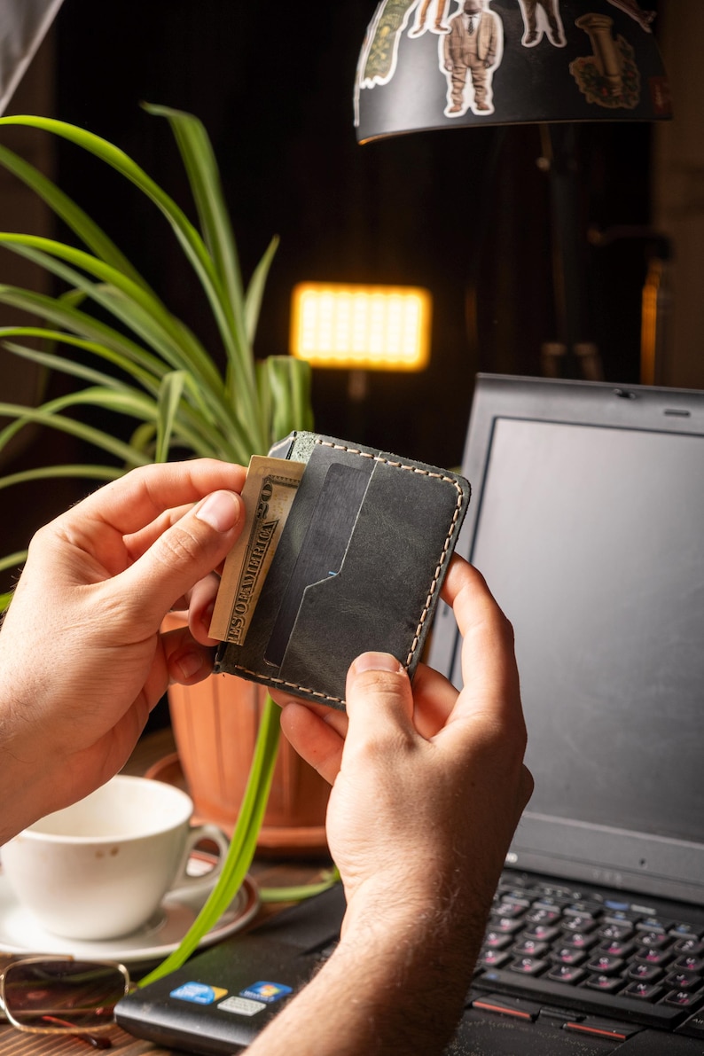 May include: A dark gray leather wallet with white stitching, holding a U.S. dollar bill. The wallet has multiple card slots and is held in front of a laptop. The background includes a plant and a lamp.