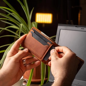 May include: A brown leather wallet with multiple card slots is being held open, revealing dark cards. The wallet is a warm, medium brown color and appears to be made of high-quality leather. The background includes a laptop and a green plant.